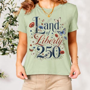 May include: A light sage green short-sleeve t-shirt featuring a graphic print with the text Land of Liberty 250 in dark blue and red lettering. The design is surrounded by illustrations of red poppies, blue cornflowers, white daisies, and butterflies.