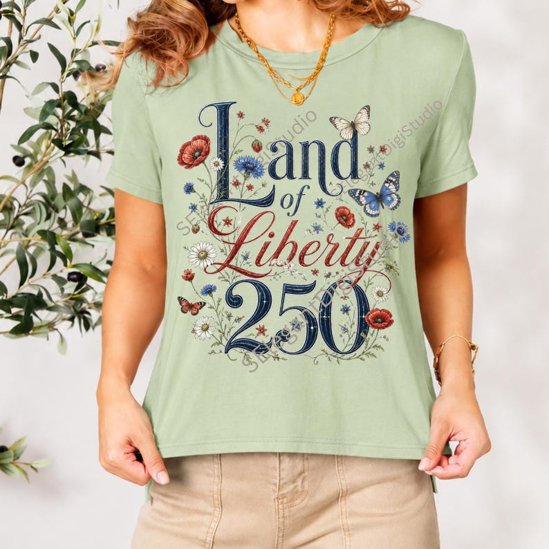 May include: A light sage green short-sleeve t-shirt featuring a graphic print with the text Land of Liberty 250 in dark blue and red lettering. The design is surrounded by illustrations of red poppies, blue cornflowers, white daisies, and butterflies.