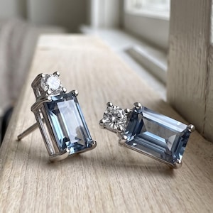 Può includere: Un paio di orecchini a lobo con gemme rettangolari blu-grigie taglio smeraldo incastonate in metallo color argento. Ogni orecchino è impreziosito da una piccola pietra rotonda trasparente sfaccettata posizionata sopra la gemma principale.