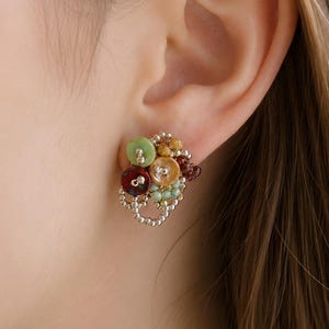 May include: A cluster earring featuring a collection of small, round, multi-colored elements in shades of green, red, yellow, and brown, accented with tiny metallic silver beads arranged in a delicate, openwork pattern that sits against the earlobe.