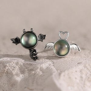 May include: A pair of silver-toned stud earrings. One earring features a devil design with horns, wings, and a tail, while the other has a heart-shaped halo and angel wings. Both earrings have a round, iridescent, dark green pearl center.