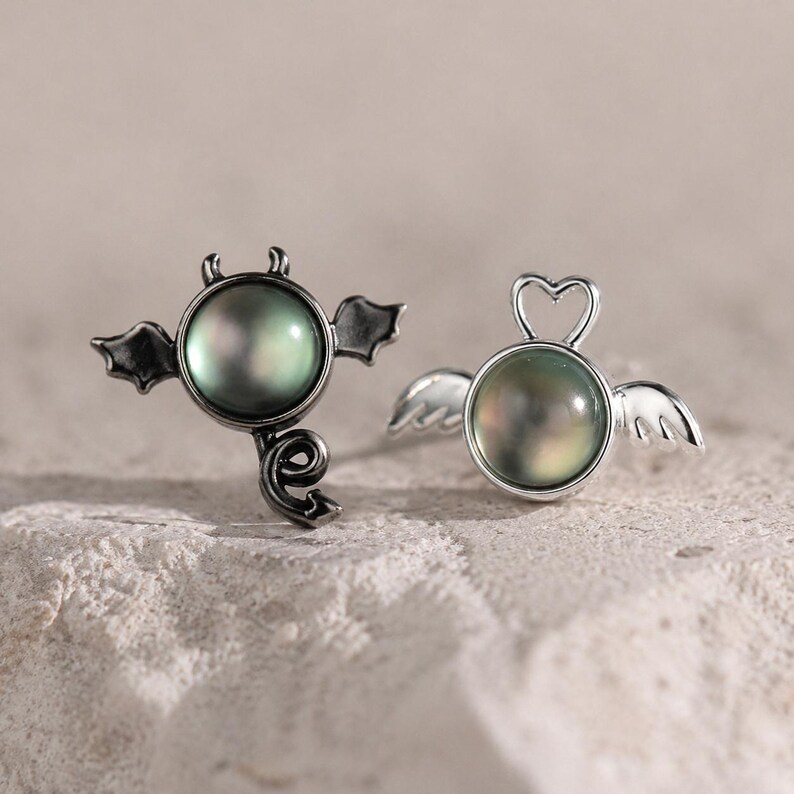 May include: A pair of silver-toned stud earrings. One earring features a devil design with horns, wings, and a tail, while the other has a heart-shaped halo and angel wings. Both earrings have a round, iridescent, dark green pearl center.