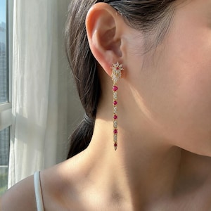 May include: A gold-toned earring with a floral design at the top and a long, dangling chain. The chain is adorned with alternating red and clear gemstones. The earring is worn on the ear.