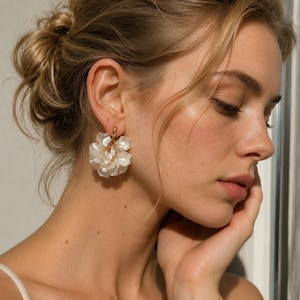 May include: A woman wearing white flower-shaped earrings. The earrings have a gold-coloured post and are made of multiple layers of white petals. The woman has blonde hair and is looking down. The earrings are a statement piece.