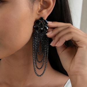 May include: A long, dangling earring featuring a cluster of black oval-shaped stones at the top, from which four cascading, layered chains of small, dark, faceted beads hang down in a graduated, looped design, creating a dramatic, multi-strand effect.