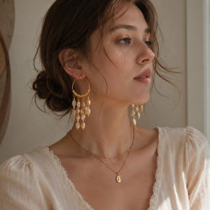 May include: Gold-tone hoop earrings with dangling pearl-like beads. A delicate gold necklace with a small pendant is also visible. The jewelry is worn with a cream-colored blouse, creating a classic and elegant look.