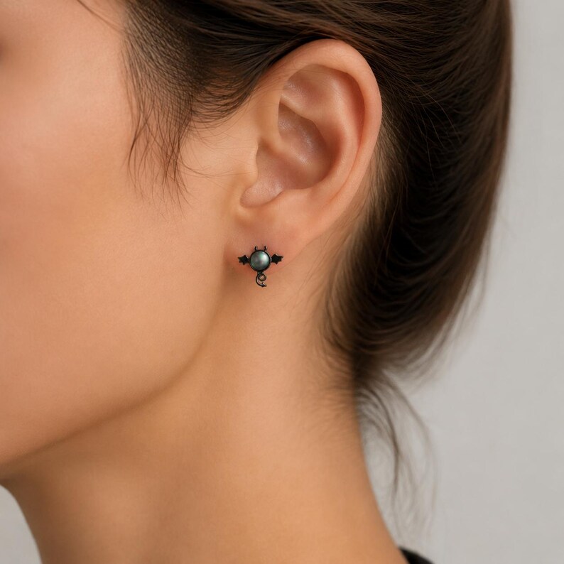 May include: A black stud earring with a bat design, featuring a dark gray pearl center. The bat has outstretched wings and a small, curled tail. The earring is worn on the ear.
