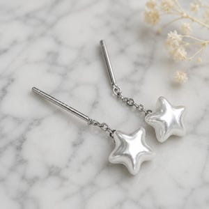 May include: A pair of silver-toned drop earrings. Each earring features a star-shaped, pearlescent charm suspended from a chain. The earrings have a post-and-clutch closure and are displayed on a marble surface.