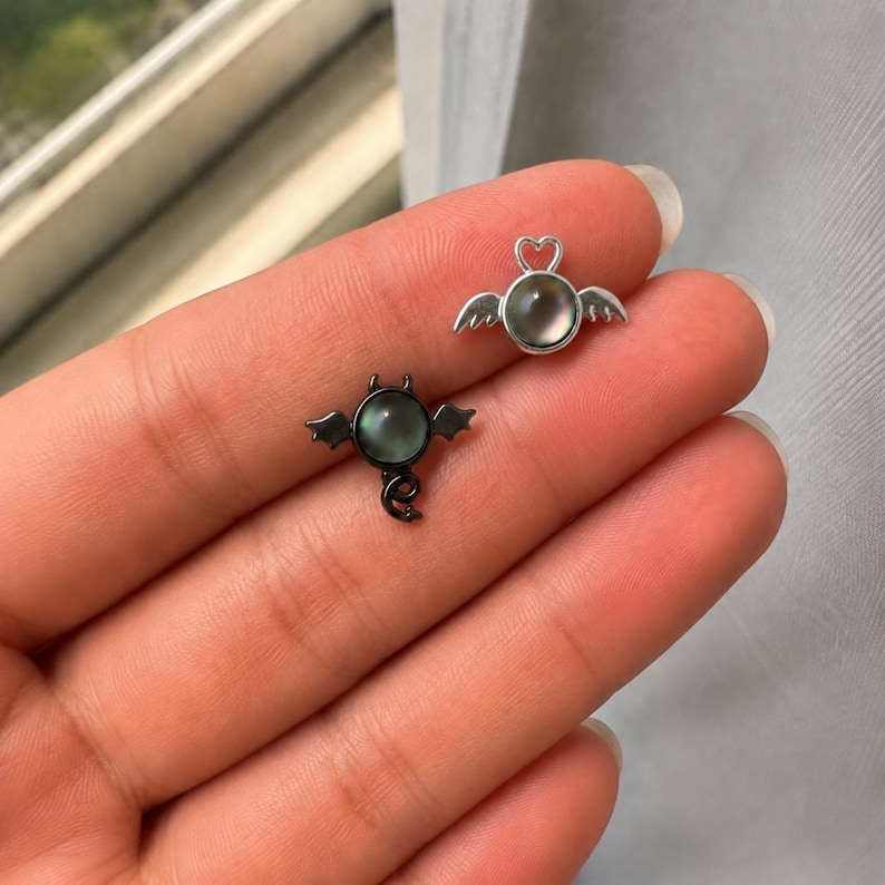 May include: Two small, decorative earrings. One is a silver angel with a heart-shaped halo and wings, the other is a black devil with horns, bat wings, and a curled tail. Both have a round, iridescent center.