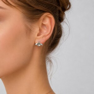 May include: Silver-toned angel wing earrings with a heart-shaped top and a round, dark-colored center stone. The earrings are small and delicate, designed to be worn in the ear. The wings are detailed with feather-like lines.
