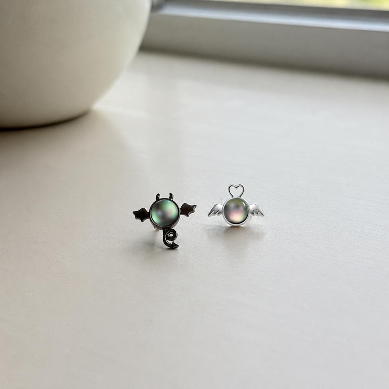 May include: A pair of unique stud earrings. One earring features a devil design with black wings and a swirling tail, while the other has an angel design with silver wings and a heart-shaped halo. Both earrings have a round, iridescent center.