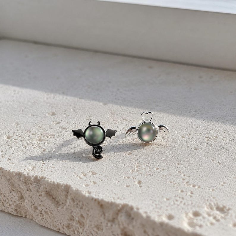 May include: Two stud earrings. One is a black bat design with a pearlescent orb, the other is a silver angel design with a pearlescent orb. The earrings are on a textured, off-white surface.