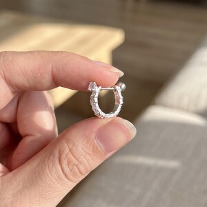 May include: A small silver-toned hoop earring featuring a hinged closure mechanism with a smooth spherical end. The outer surface of the hoop is embellished with a series of small, clear, round-cut stones set along the curved metal frame.