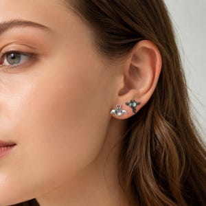 May include: A pair of earrings, one with a heart-shaped design and wings, the other with a dark, round gem and wings. The earrings are silver-toned and are displayed on an ear.