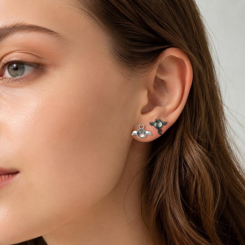 May include: A pair of earrings, one with a heart-shaped design and wings, the other with a dark, round gem and wings. The earrings are silver-toned and are displayed on an ear.