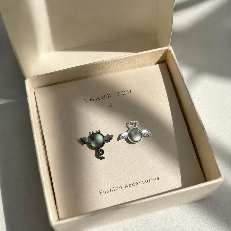 May include: A pair of silver-toned stud earrings, one with a devilish design and the other with angel wings, presented in a gift box. The earrings feature iridescent, round center stones. The card inside the box reads "THANK YOU" and "Fashion Accessories".