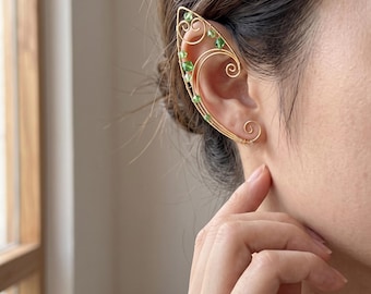 Könnte beinhalten: Eine goldfarbene Ohrklemme aus Draht in Form eines länglichen Blattes oder Flügels, mit zarten, geschwungenen Drahtmustern und kleinen, facettierten grünen Perlen am Außenrand, die um den oberen Knorpel des Ohrs gelegt wird.