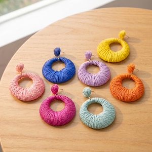 May include: A collection of seven circular earrings featuring a woven raffia texture. The earrings are available in a variety of vibrant colors including yellow, blue, lavender, orange, pink, magenta, and light teal, each with a small stud top.