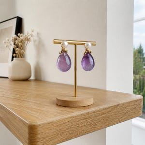 Könnte beinhalten: Ein Paar Amethyst-Tropfenohrringe mit goldfarbenem Steg und Perlenakzent. Die Ohrringe werden auf einem goldfarbenen Ständer mit Holzsockel präsentiert. Die Ohrringe haben eine tiefviolette Farbe.