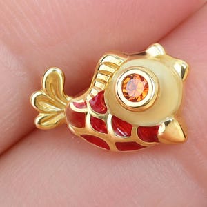 May include: A gold-toned fish-shaped charm featuring a bright orange circular gemstone set as the eye. The body of the fish is decorated with a glossy red enamel pattern that fills the spaces between the raised gold-colored metal scales.