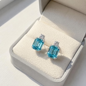 May include: A pair of stud earrings featuring emerald-cut, bright blue gemstones set in silver-toned metal. Each earring is accented with a single, small, round, clear, faceted stone positioned directly above the larger blue rectangular gem.