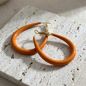 May include: A pair of teardrop-shaped hoop earrings featuring a smooth, glossy orange enamel finish. The earrings are constructed with a gold-toned metal post and a hinged clasp mechanism, creating a continuous, rounded silhouette for the jewelry.