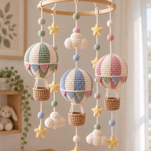 May include: A nursery mobile featuring three crocheted hot air balloons in sage green, dusty blue, and muted pink, each with a brown basket. The mobile includes crocheted clouds and yellow stars suspended from a circular wooden frame by white cords.