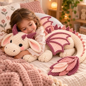 May include: A large, handmade crochet dragon plush toy featuring a cream-coloured body with soft pink and deep purple accents on the wings, horns, and tail. The stuffed creature has large black safety eyes, a rounded snout, and a long, segmented tail.