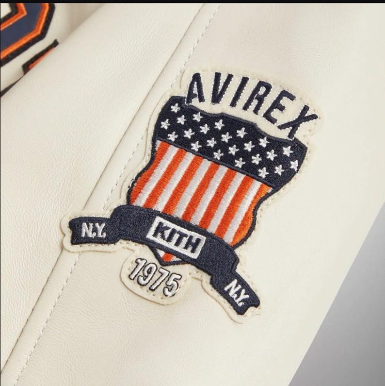May include: An off-white leather jacket sleeve featuring an embroidered patch. The patch displays the text AVIREX above a shield with stars and stripes, a banner reading KITH with N.Y. on both sides, and the year 1975 stitched in white thread.