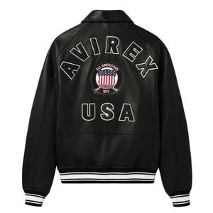 May include: A black leather varsity jacket featuring the white embroidered text AVIREX in an arc across the upper back. Below this is a circular patch with the American flag and the text ALL AMERICANS ESTABLISHED 1975. The word USA is embroidered in white below the patch, with striped black and white ribbed cuffs and hem.