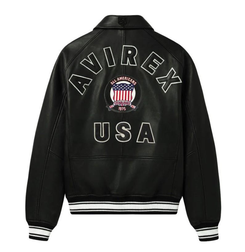 May include: A black leather varsity jacket featuring the white embroidered text AVIREX in an arc across the upper back. Below this is a circular patch with the American flag and the text ALL AMERICANS ESTABLISHED 1975. The word USA is embroidered in white below the patch, with striped black and white ribbed cuffs and hem.