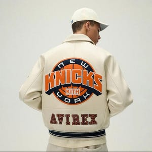 May include: An off-white varsity jacket featuring a large circular New York Knicks basketball logo on the back with orange and navy blue text. The word KITH is centered on the basketball graphic, and the word AVIREX is printed in dark red below.