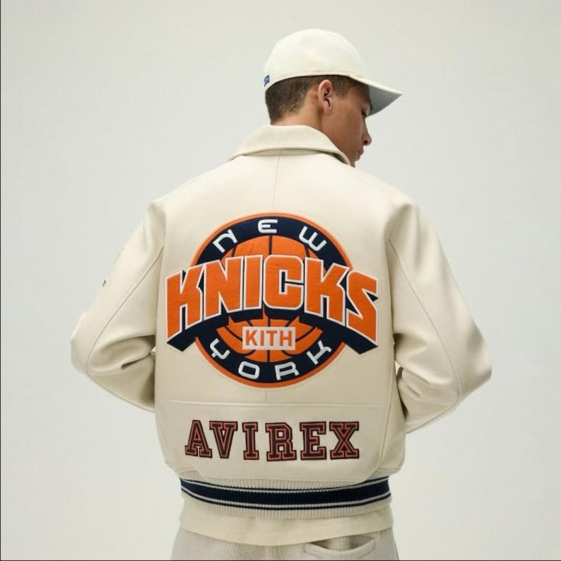 May include: An off-white varsity jacket featuring a large circular New York Knicks basketball logo on the back with orange and navy blue text. The word KITH is centered on the basketball graphic, and the word AVIREX is printed in dark red below.