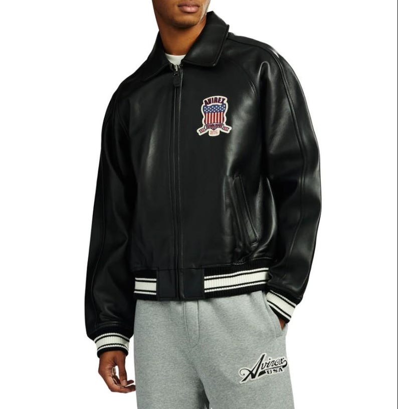 May include: A black leather jacket with a fold-down collar, a full-length front zipper, and ribbed cuffs and hem featuring white stripes. A patch on the chest displays the text AVIREX with an American flag graphic and the words ESTABLISHED 1975.