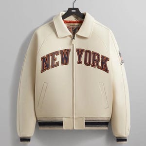 May include: A cream-colored varsity jacket featuring a full-length front zipper, a pointed collar, and ribbed cuffs and hem with navy and white stripes. The text NEW YORK is embroidered across the chest in navy blue with a dark orange outline.