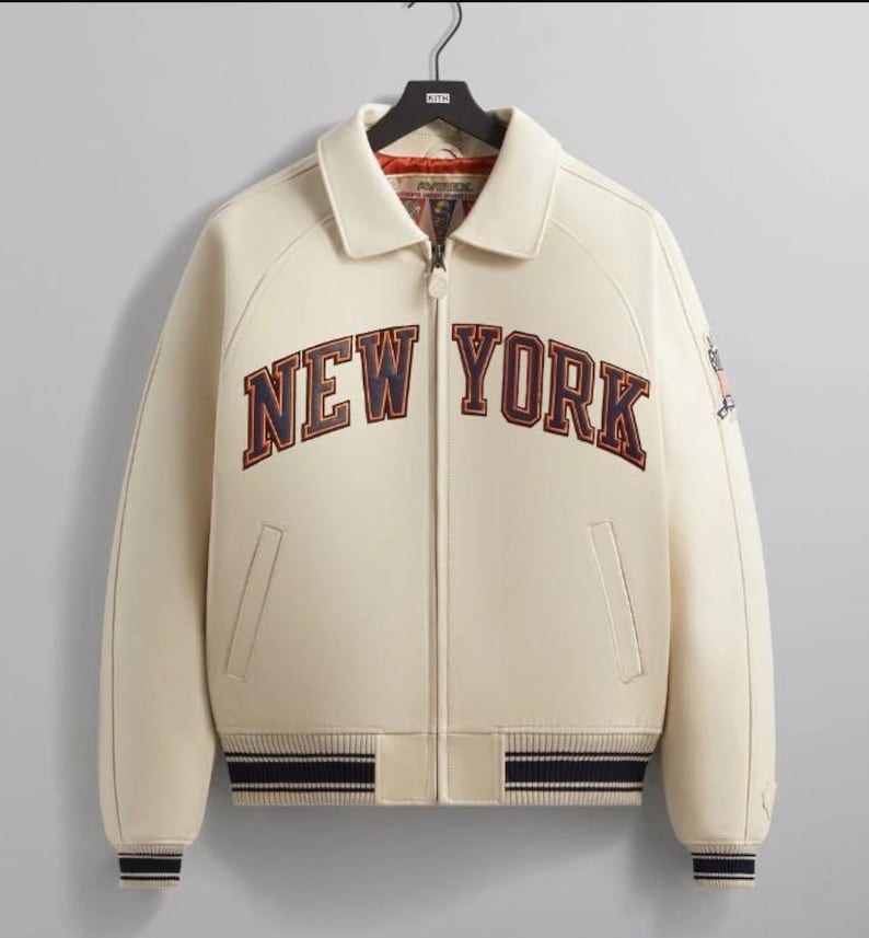 May include: A cream-colored varsity jacket featuring a full-length front zipper, a pointed collar, and ribbed cuffs and hem with navy and white stripes. The text NEW YORK is embroidered across the chest in navy blue with a dark orange outline.