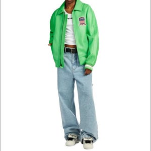May include: A bright green zip-up jacket with a collar and a small patch featuring a shield design on the chest, worn over a white tank top. The outfit includes light blue denim jeans with a black belt and white sneakers with black accents.