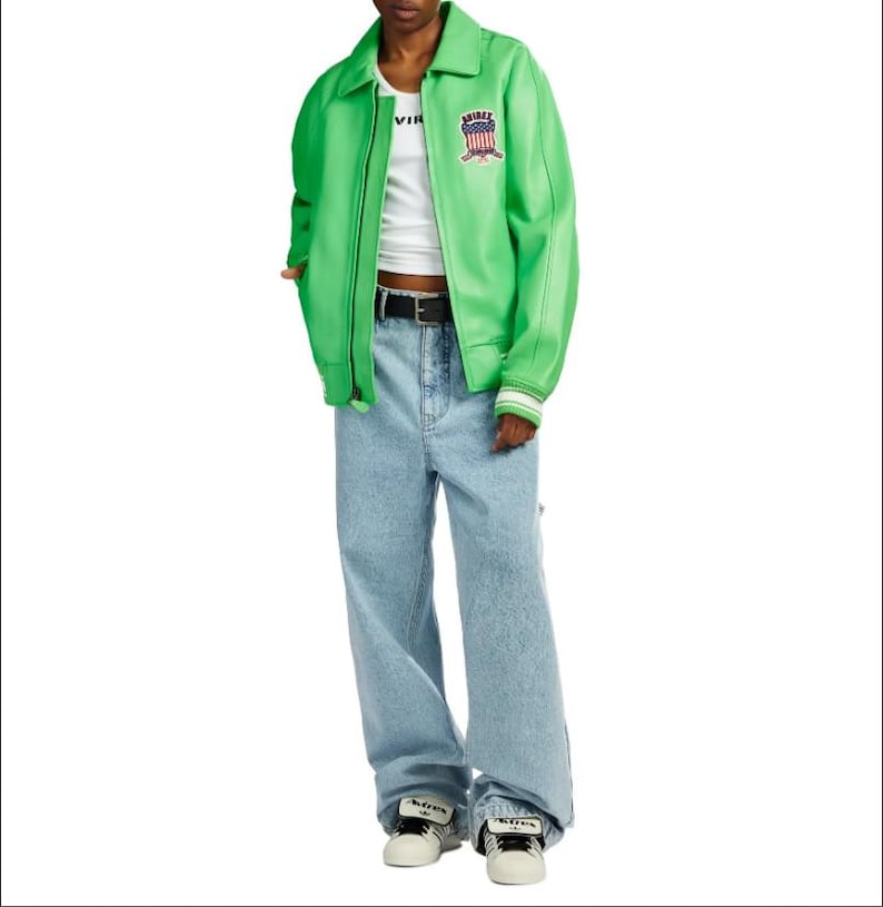 May include: A bright green zip-up jacket with a collar and a small patch featuring a shield design on the chest, worn over a white tank top. The outfit includes light blue denim jeans with a black belt and white sneakers with black accents.