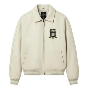 May include: An off-white leather jacket featuring a classic collar, a full-length front zipper, and ribbed cuffs and hem. A black and white embroidered patch on the left chest displays the text AVIREX, ESTABLISHED, and 1975 with a shield graphic.