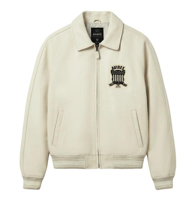 May include: An off-white leather jacket featuring a classic collar, a full-length front zipper, and ribbed cuffs and hem. A black and white embroidered patch on the left chest displays the text AVIREX, ESTABLISHED, and 1975 with a shield graphic.