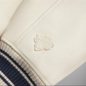 May include: An off-white jacket sleeve made of smooth material featuring a tonal embroidered patch of a basketball and a building silhouette. The sleeve ends in a ribbed knit cuff with alternating dark navy blue and light beige horizontal stripes.