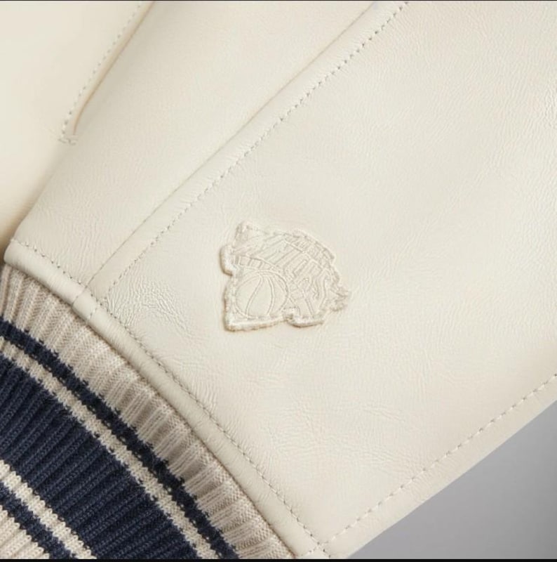 May include: An off-white jacket sleeve made of smooth material featuring a tonal embroidered patch of a basketball and a building silhouette. The sleeve ends in a ribbed knit cuff with alternating dark navy blue and light beige horizontal stripes.