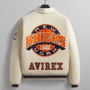 May include: A cream-colored varsity jacket featuring a large embroidered New York Knicks logo on the back with the text KITH inside. The word AVIREX is embroidered in dark lettering below the logo. The jacket has dark striped ribbed cuffs and hem.