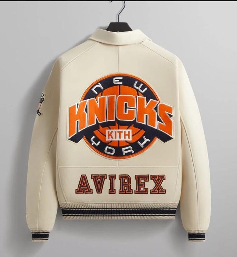 May include: A cream-colored varsity jacket featuring a large embroidered New York Knicks logo on the back with the text KITH inside. The word AVIREX is embroidered in dark lettering below the logo. The jacket has dark striped ribbed cuffs and hem.