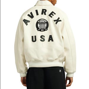 May include: An off-white leather varsity jacket featuring black lettering that reads AVIREX and USA on the back. A circular black patch with the text ALL AMERICANS ESTABLISHED 1975 and a flag graphic is centered between the words. The jacket has ribbed cuffs.