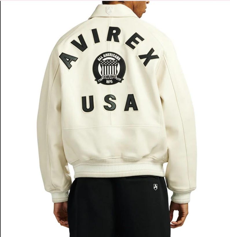 May include: An off-white leather varsity jacket featuring black lettering that reads AVIREX and USA on the back. A circular black patch with the text ALL AMERICANS ESTABLISHED 1975 and a flag graphic is centered between the words. The jacket has ribbed cuffs.
