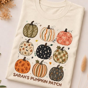 May include: A cream-colored t-shirt featuring a graphic print of nine pumpkins with various patterns including leaves, stars, stripes, and bows. The text "SARAH'S PUMPKIN PATCH" is printed in brown at the bottom of the design, which also includes small stars and hearts.