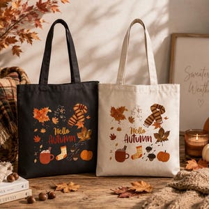 May include: Two canvas tote bags, one black and one off-white, each featuring a printed design of autumn leaves, a striped scarf, a mug, a sock, and a pumpkin with the text Hello Autumn. The bags have two shoulder straps and a rectangular shape.