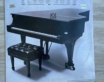 May include: A black grand piano with a black piano bench. The piano has a white background and the text "Elton John Here And There" is printed on the top of the album cover.