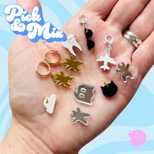 May include: A collection of small acrylic charms and earrings featuring various shapes including a white tooth, black sunglasses, a white airplane, a gold gingerbread person, a white cloud, a silver ghost, a black cat head, and a silver gingerbread person.